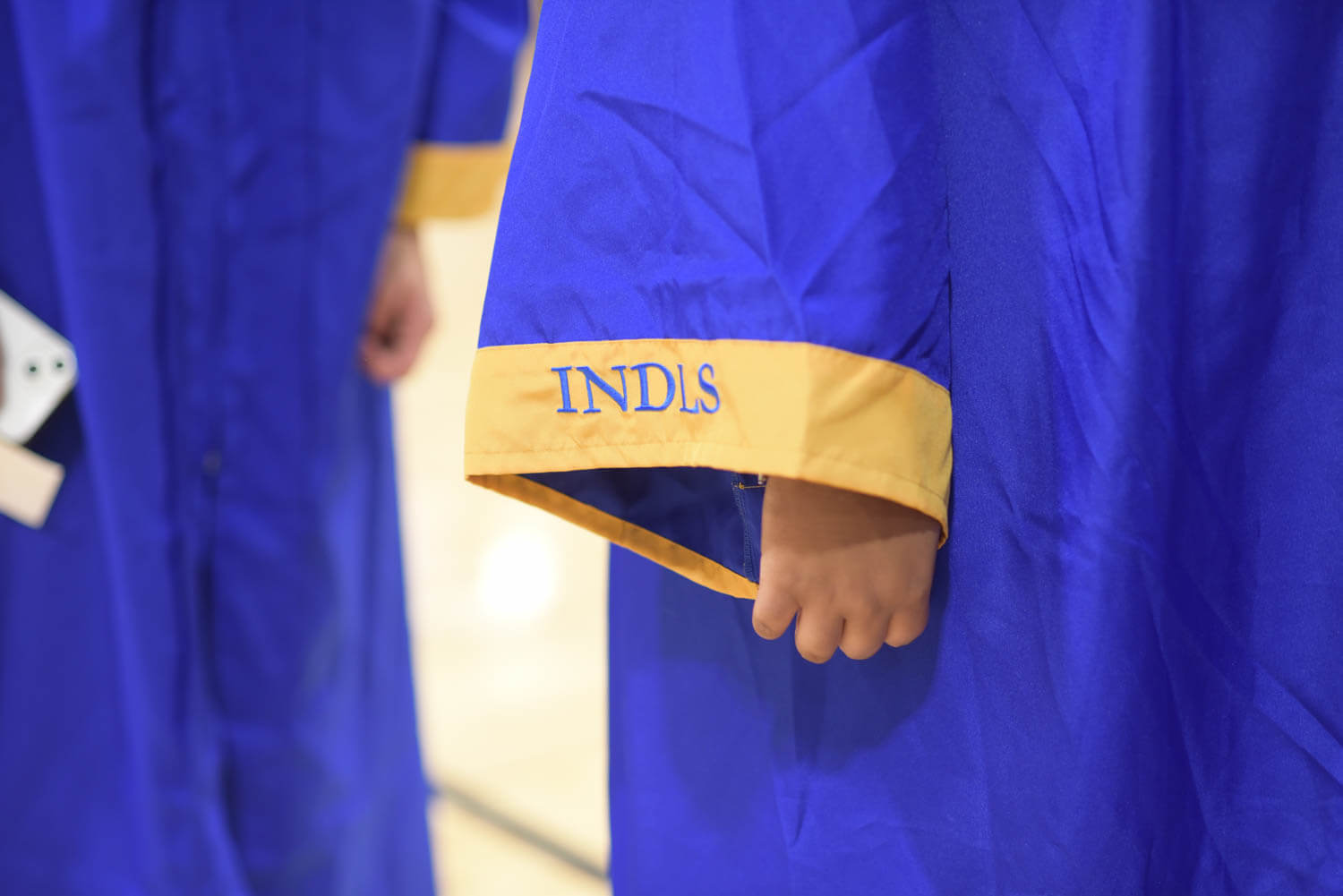 Close-up of a child’s hand holding a blue graduation gown with a gold hem and "INDLS" embroidered.