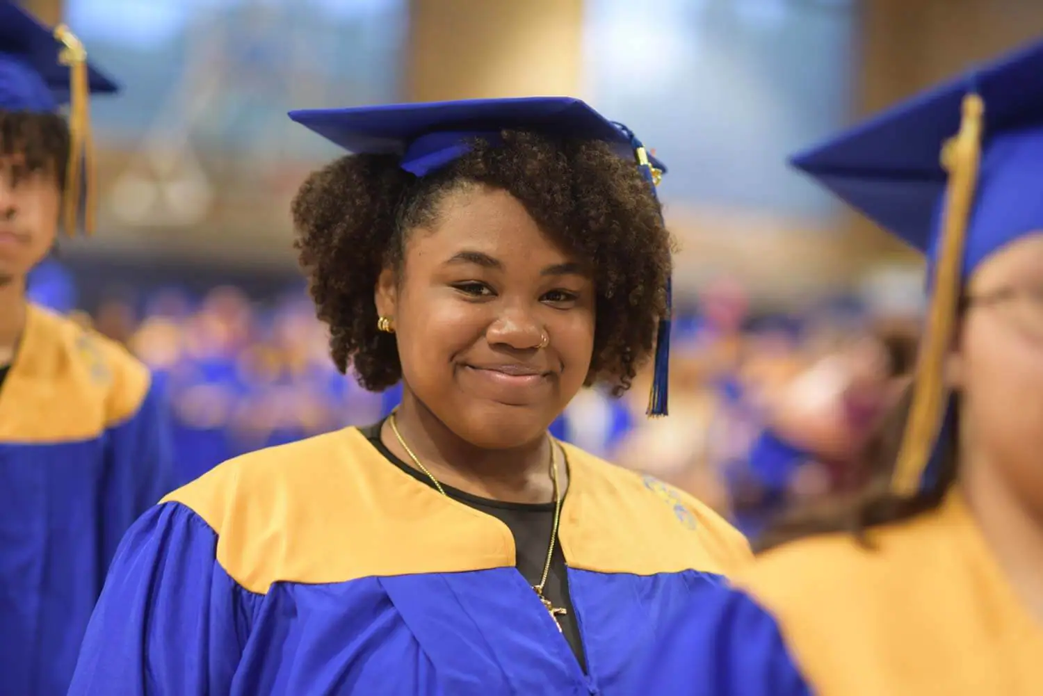 A graduate wearing a blue gown and cap with a gold sash smiles confidently at the camera.