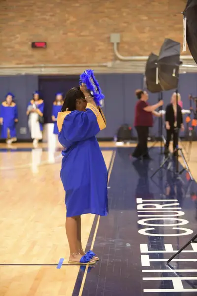 A graduate in a blue gown and cap adjusts her tassel on a gymnasium floor.