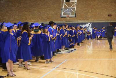 Graduates in blue and gold gowns line a gymnasium, poised with excitement.