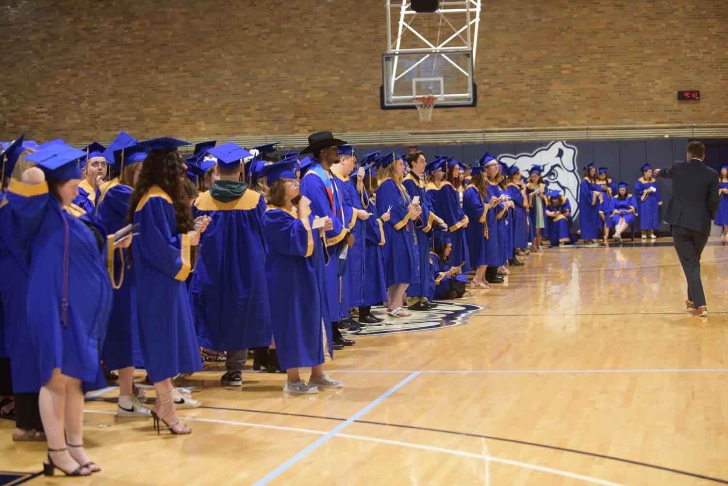 Graduates in blue and gold gowns line a gymnasium, poised with excitement.