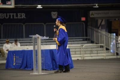 A graduate in a blue cap and gown speaks at a clear podium on a stage.