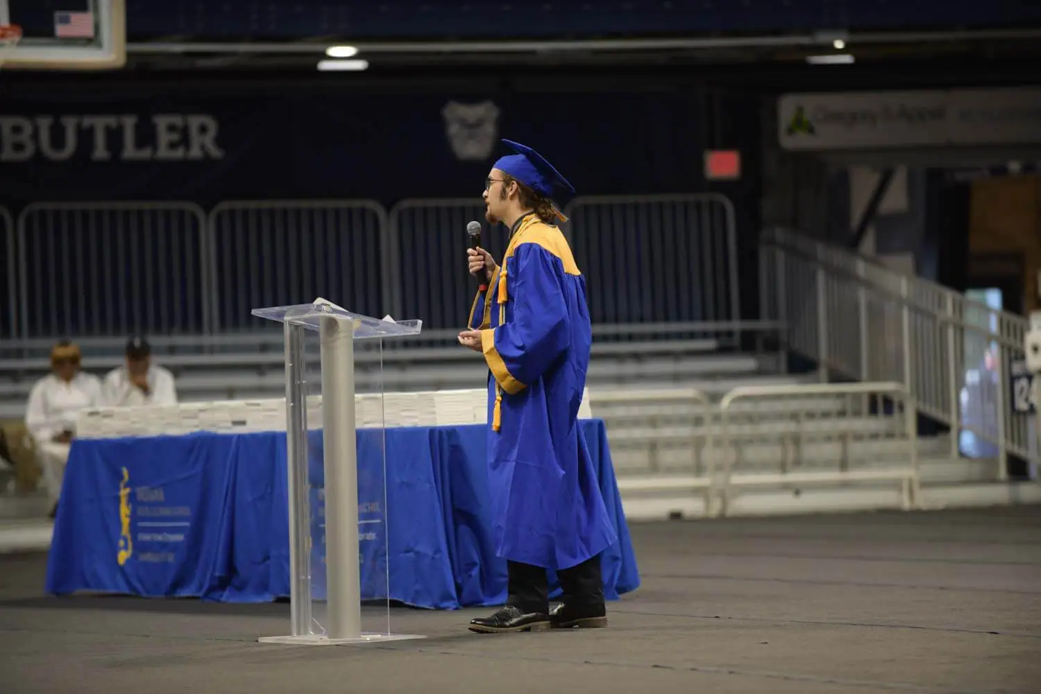 A graduate in a blue cap and gown speaks at a clear podium on a stage.