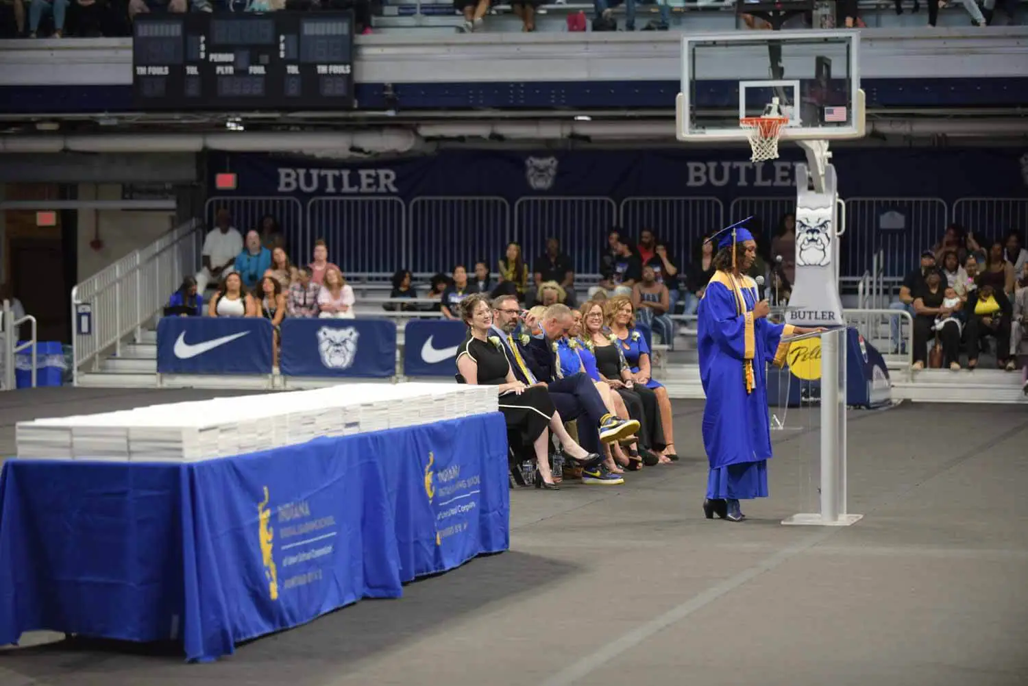 A graduate in a blue cap and gown speaks at a podium on a gymnasium floor.