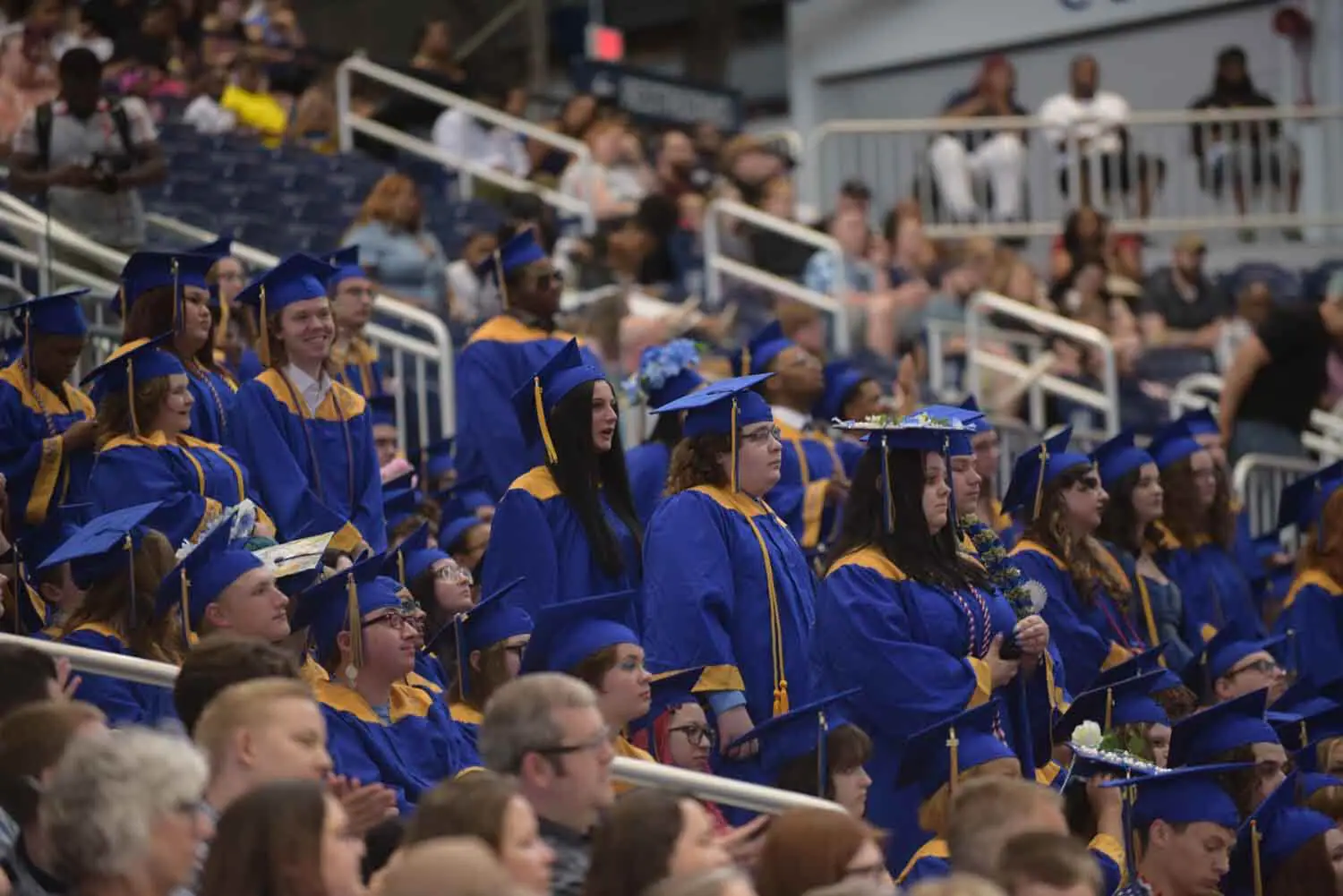 Graduates in blue caps and gowns stand and sit in a crowded stadium, some decorated with flowers.