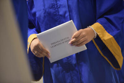 A person in a blue graduation gown with gold trim holds a diploma