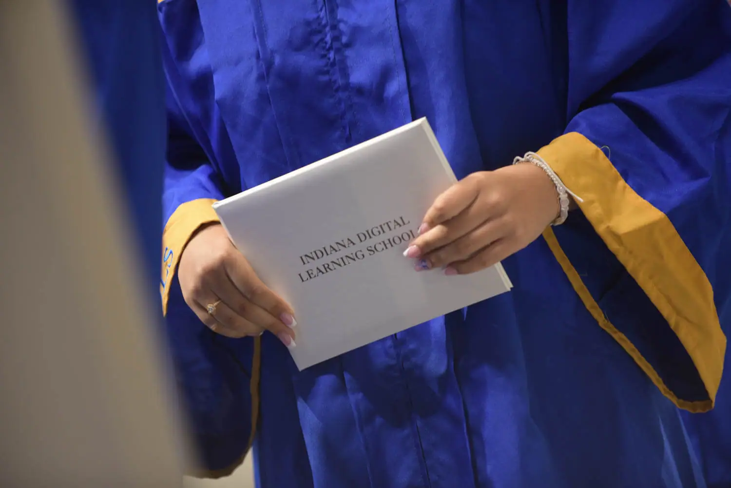 A person in a blue graduation gown with gold trim holds a diploma