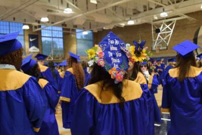 Graduates in blue gowns with yellow stoles gather in a gym.