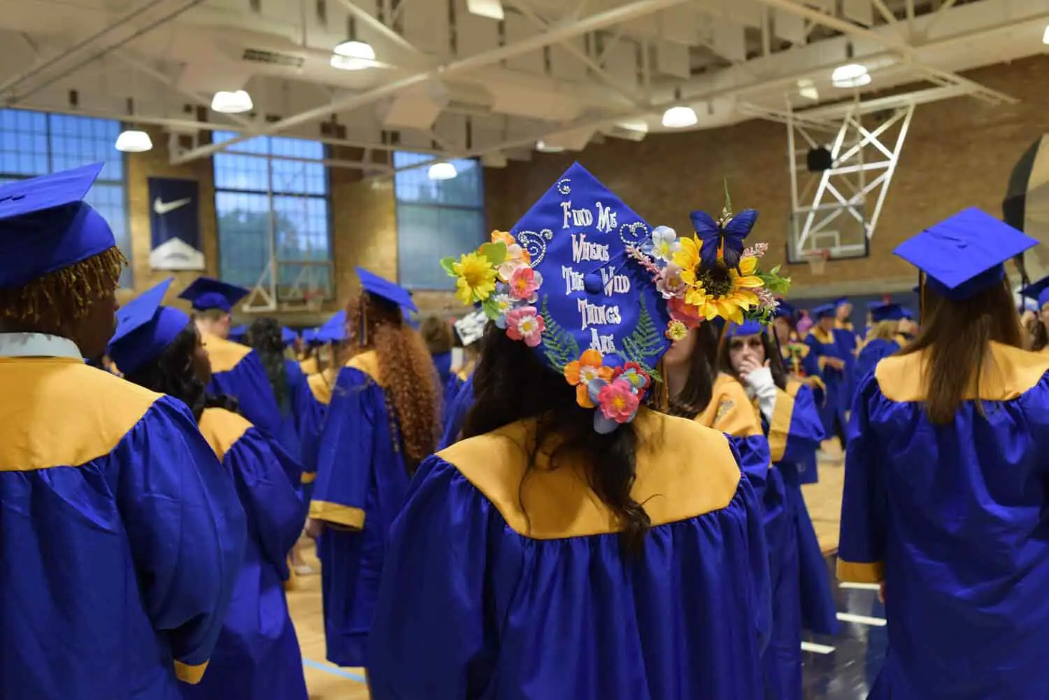 Graduates in blue gowns with yellow stoles gather in a gym.