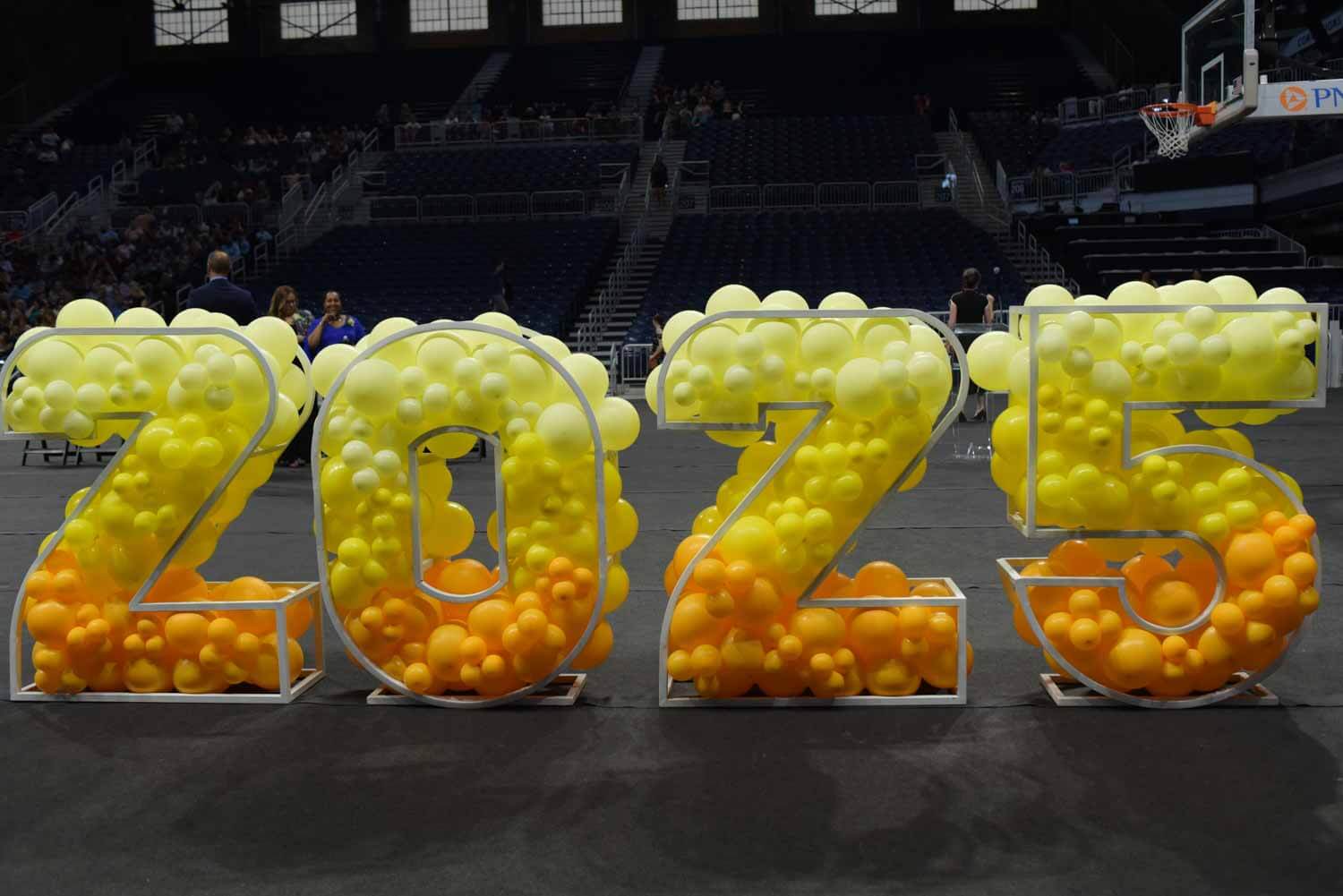 Large balloon numbers "2025" in yellow and orange gradient stand in an indoor arena.