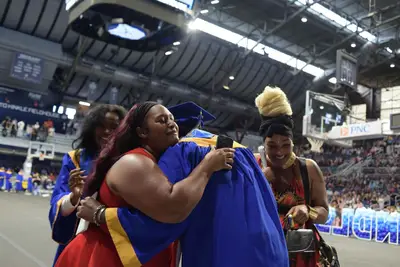 Graduate in blue cap and gown hugs a woman in red, surrounded by joyful supporters in an arena.