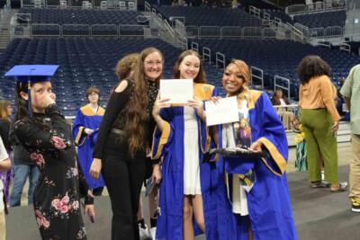 Graduates in blue and gold gowns joyfully hold diplomas, posing with a smiling woman, amidst a backdrop of a large indoor stadium.