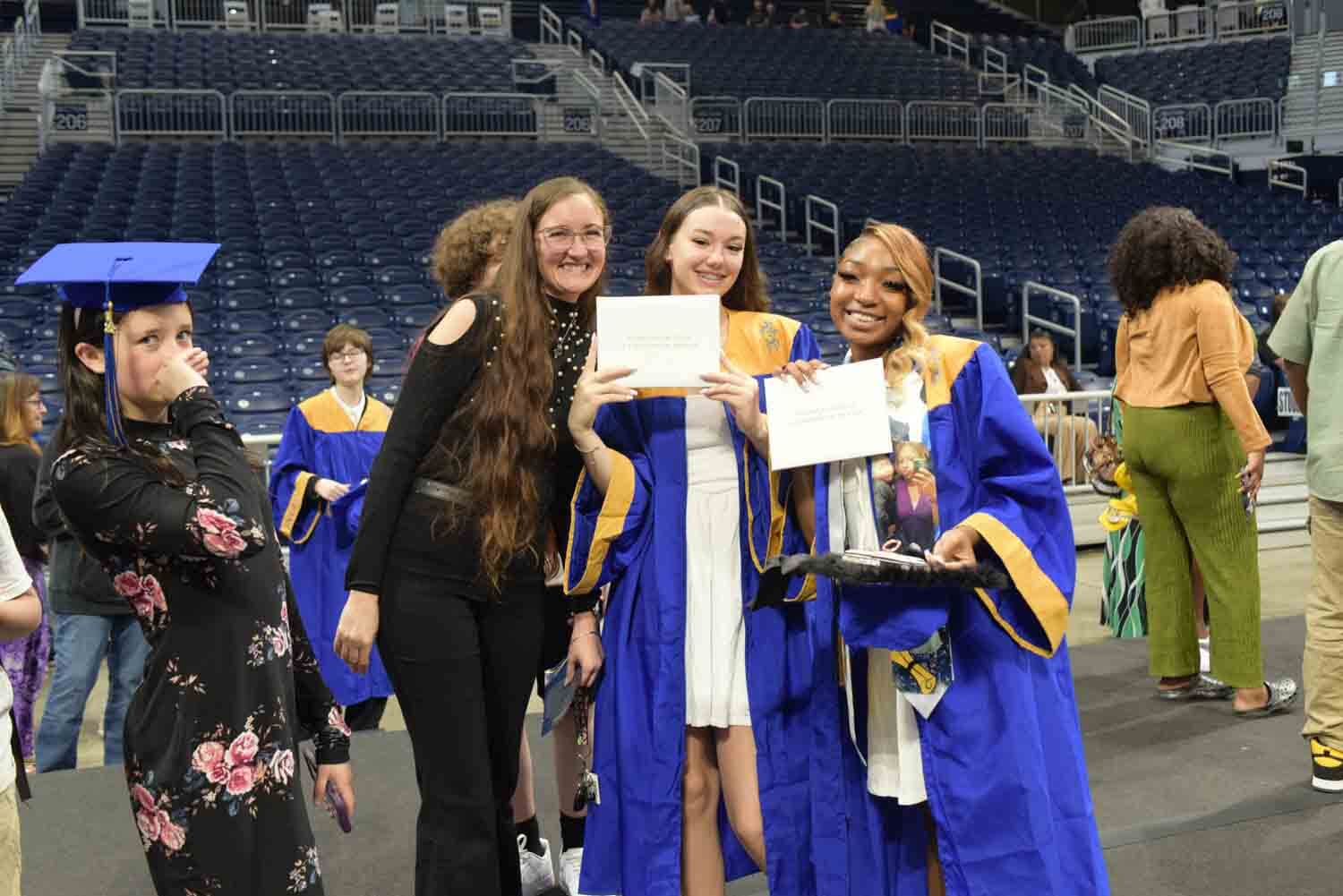 Graduates in blue and gold gowns joyfully hold diplomas, posing with a smiling woman, amidst a backdrop of a large indoor stadium.
